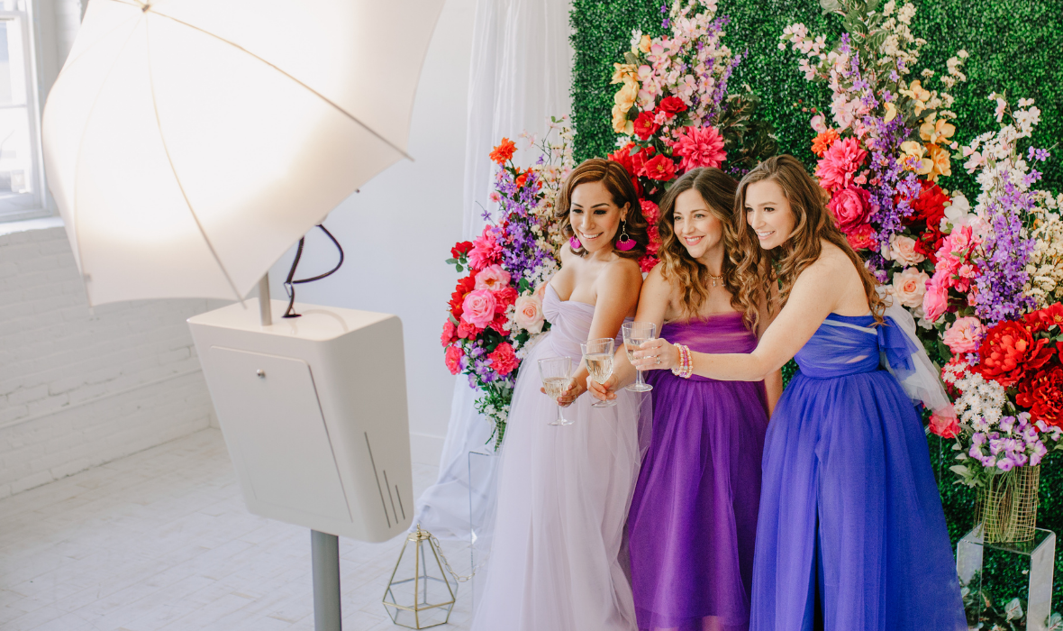 Three women taking a photo in front of flower wall wearing tulle dresses by the lotus bloom co.